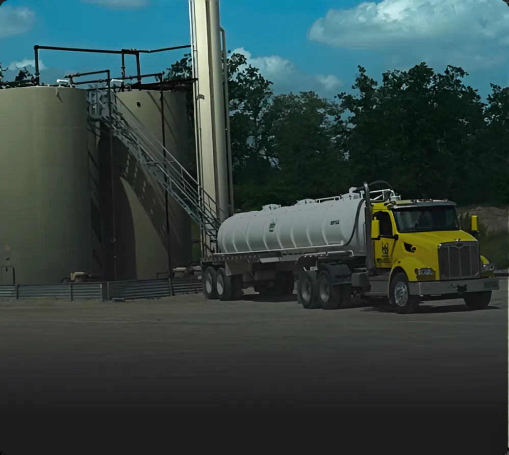 A yellow Wrangler tractor and steel tank trailer beside storage tanks